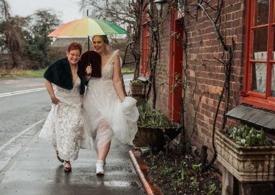 Two brides walking together under a rainbow patterned umbrella during a rainy but relaxed wedding in Bridgnorth, Shropshire